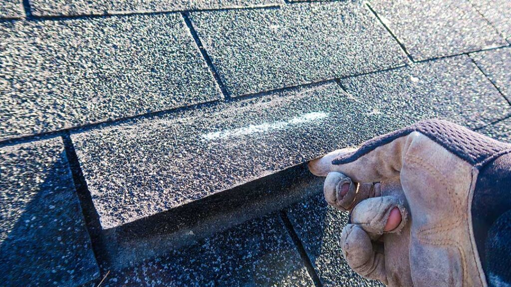 A person holding several roof shingle samples against a home's siding in natural daylight.